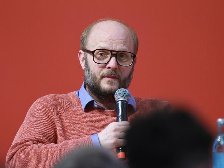 David Schraven sitzt vor einer roten Wand, hält ein Mikrofon in der Hand und scheint mit dem Publikum zu sprechen