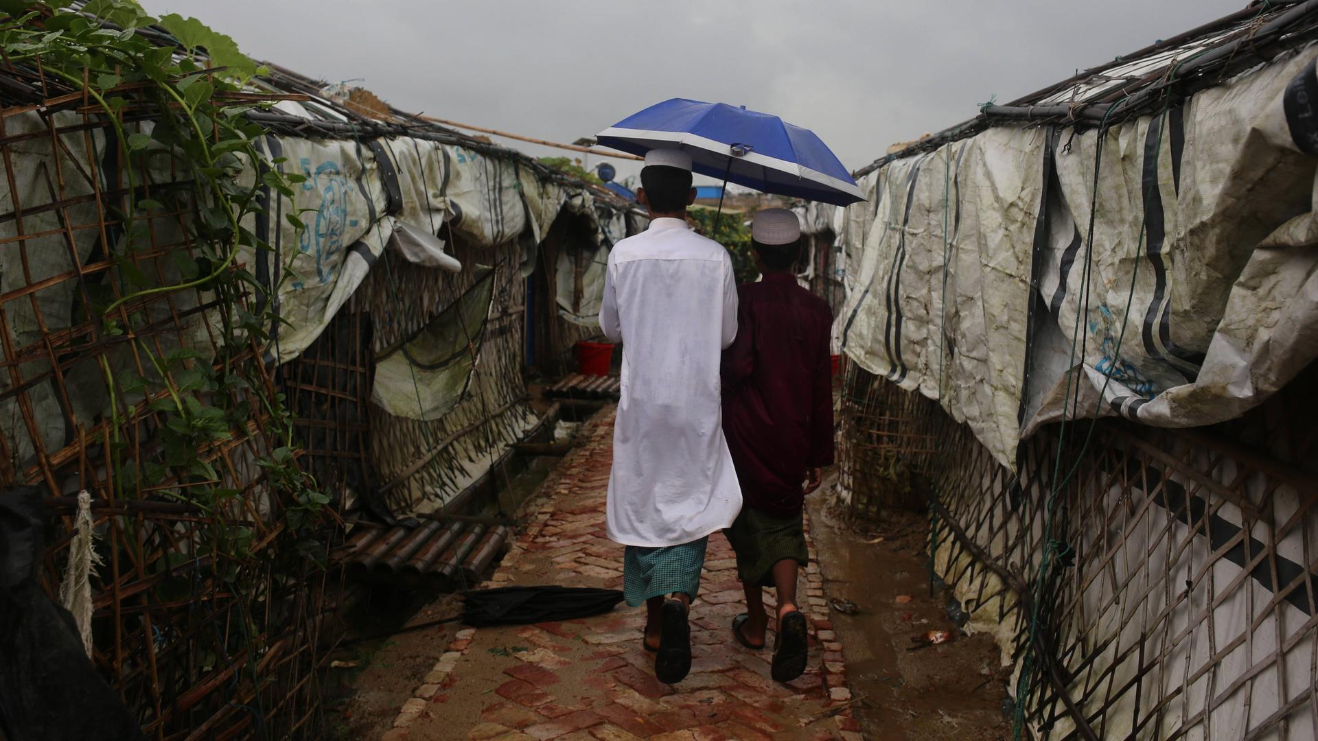 Zwei junge Männer in langen Gewändern laufen unter einem Regenschirm eine unbefestigte Straße im Camp Kutupalong entlang. Die flachen Gebäude links und recht davon bestehen aus Ästen und Planen.