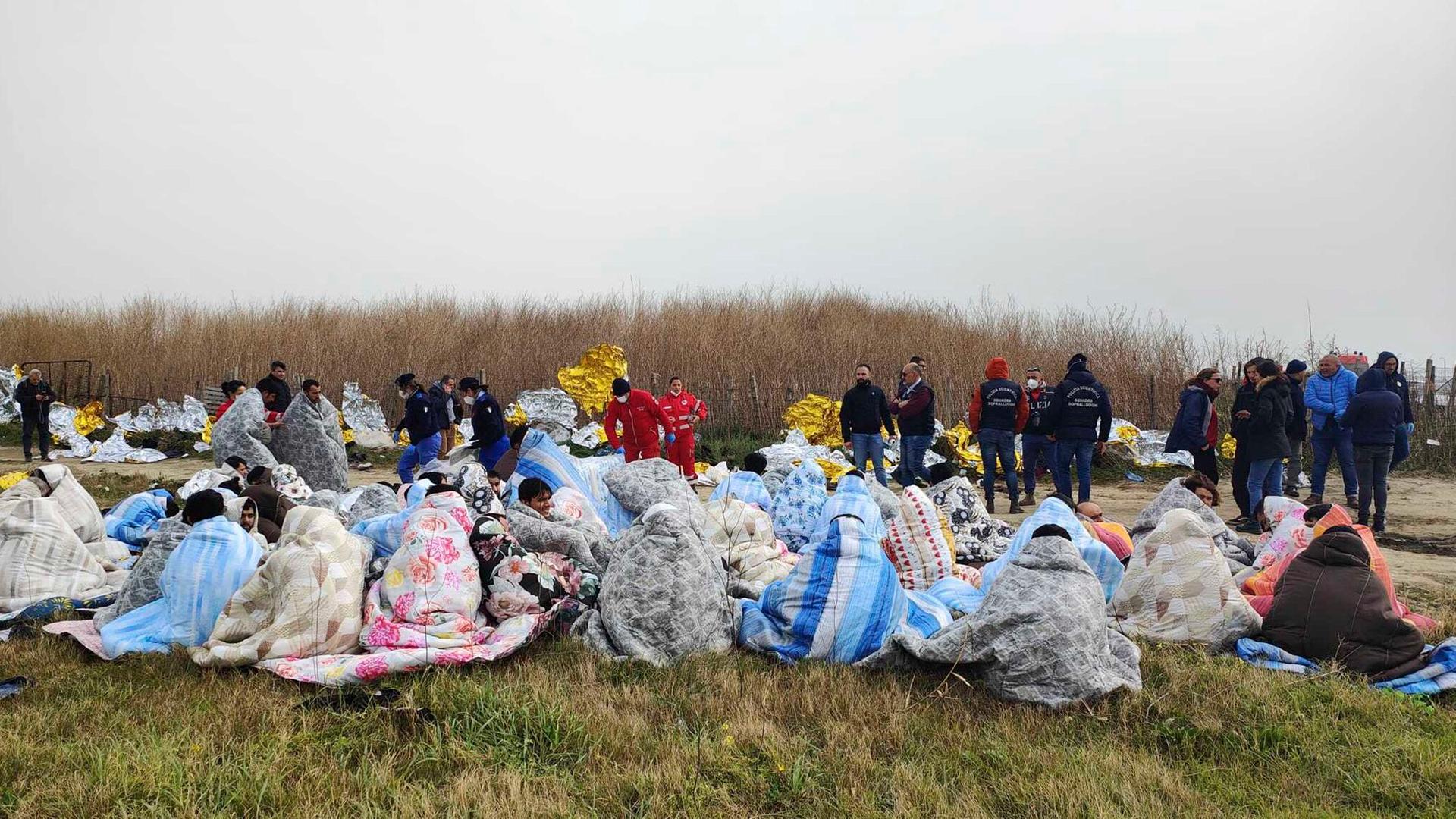 Italien, Cutro: Gerettete Migranten sitzen in Decken gehüllt an einem Strand.