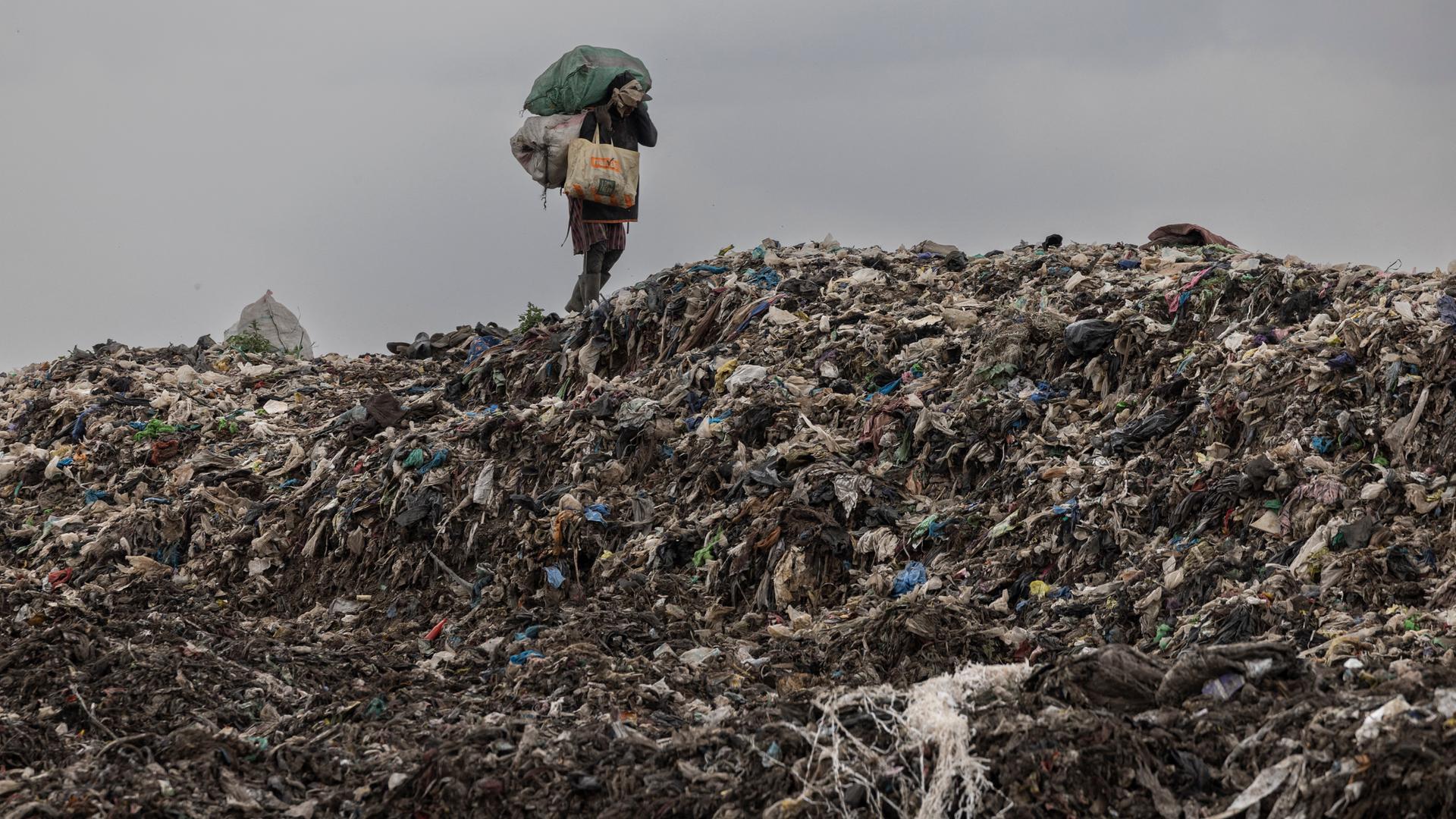 Eine Person in improvisierter Schutzkleidung läuft auf einem Müllberg entlang und sammelt recyclebaren Müll.