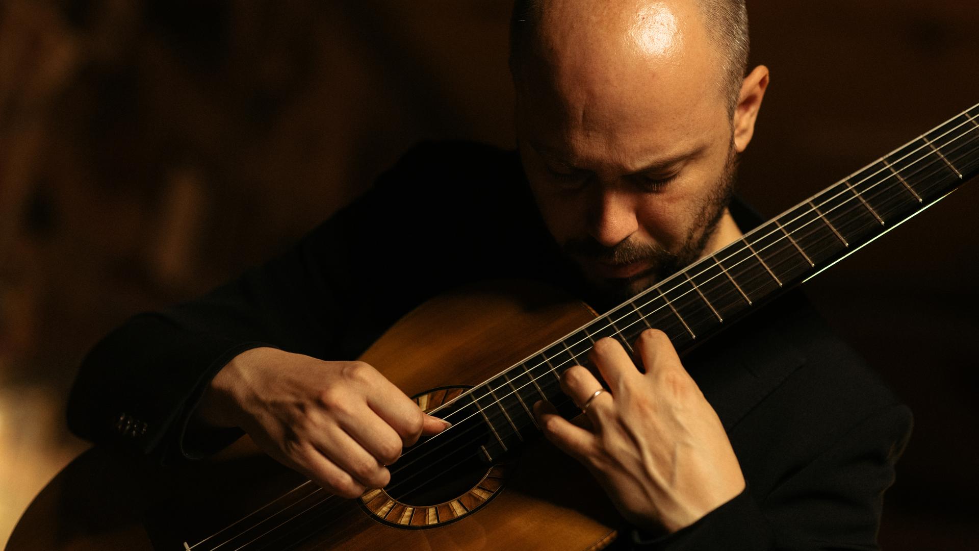 Ein Mann spielt Gitarre, er beugt seinen Kopf dabei über das Instrument. Das Foto ist in dunklen Tönen gehalten.