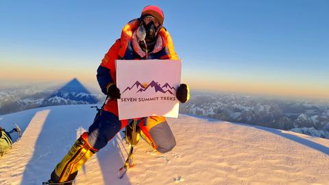 Ein Bergsteiger auf einem schneebedeckten Gipfel hält ein Plakat mit der Aufschrift "Seven Summit Treks".