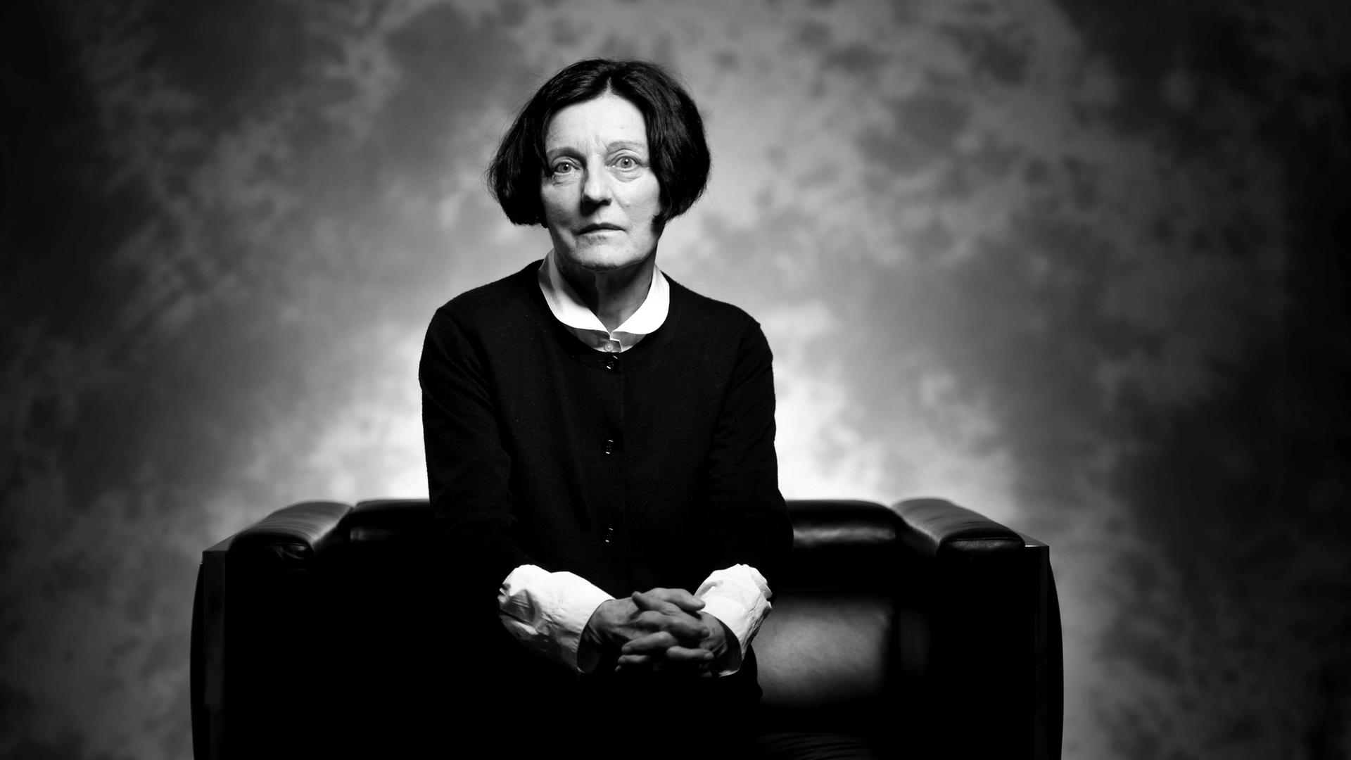 Schwarz-Weiß-Porträt der Schriftstellerin und Literatur-Nobelpreis-Trägerin Herta Müller, die auf einem schwarzen Sofa sitzt und mit gefalteten Händen ernst in die Kamera blickt.