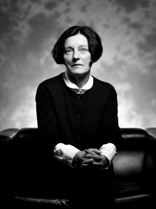 Schwarz-Weiß-Porträt der Schriftstellerin und Literatur-Nobelpreis-Trägerin Herta Müller, die auf einem schwarzen Sofa sitzt und mit gefalteten Händen ernst in die Kamera blickt.