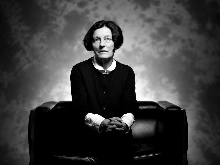 Schwarz-Weiß-Porträt der Schriftstellerin und Literatur-Nobelpreis-Trägerin Herta Müller, die auf einem schwarzen Sofa sitzt und mit gefalteten Händen ernst in die Kamera blickt.