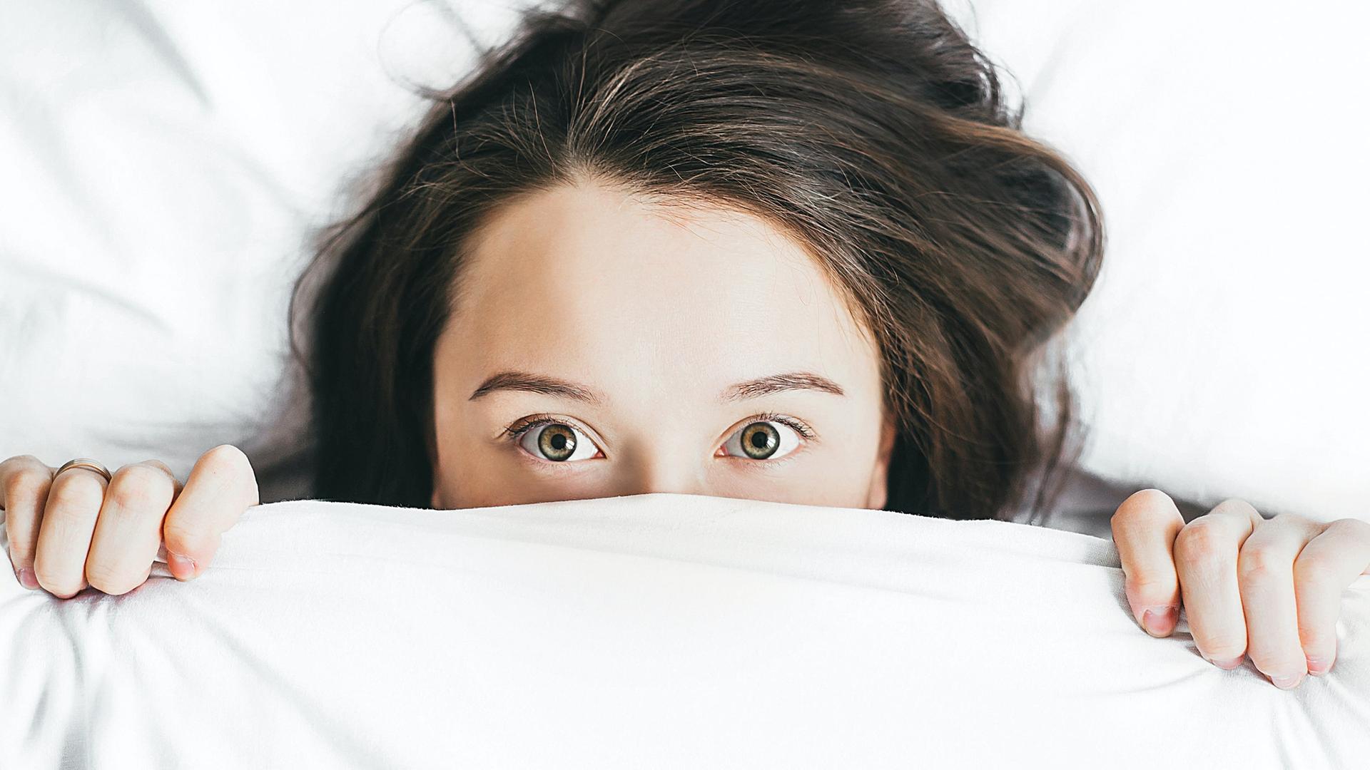 Eine Frau zieht sich die Bettdecke übers Gesicht, nur noch ihre Augen, Stirn und Haaren schauen darunter hervor. 