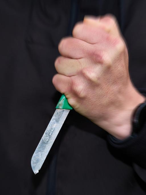 Ein schwarz gekleideter Mann hält ein Messer mit fester Faust. 