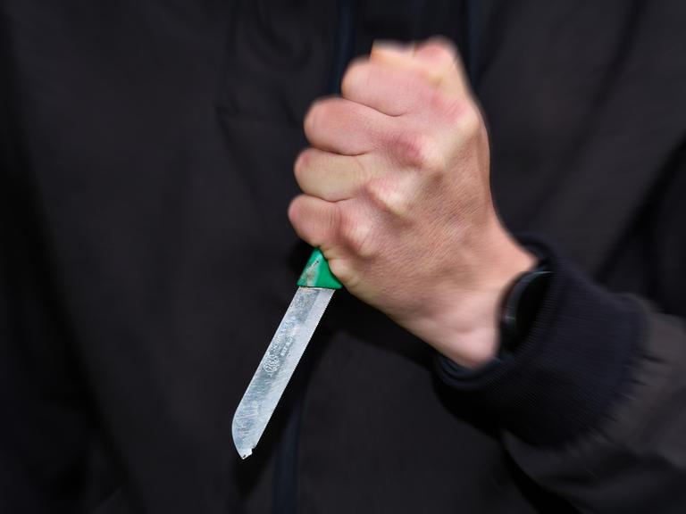 Ein schwarz gekleideter Mann hält ein Messer mit fester Faust. 