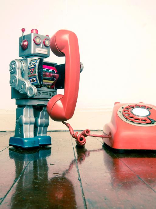 Ein silberner roter Spielzeugroboter hält den Hörer eines roten Wählscheinbentelefons, das neben im steht, in der Hand.