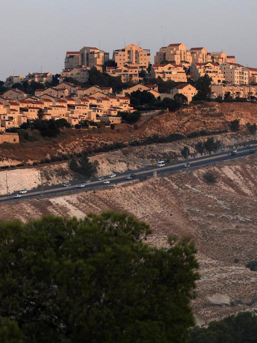 Autos fahren auf einer Straße unterhalb einer großen Siedlung moderner Häuser entlang. Es ist eine israelische Siedlung außerhalb von Jerusalem im Westjordanland.