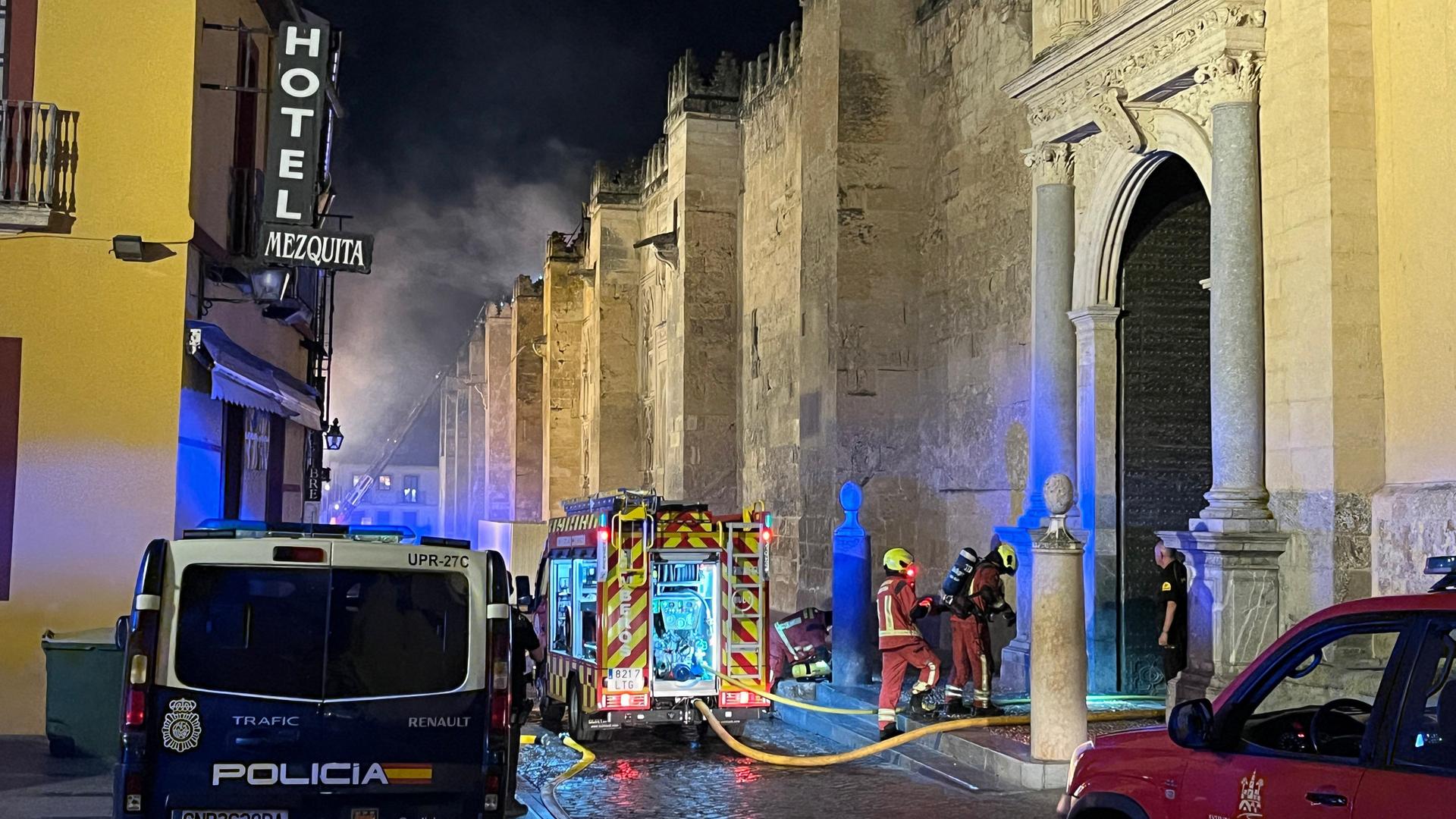 Es ist dunkel. Links ein Hotel. Rechts an der Mauer der Kirche stehen Feuerwehrfahrzeuge und Feuerwehrmänner, die einen Schlauch ins innere des Gebäudes verlegt haben. Im Hintergrund steigt Rauch auf, 