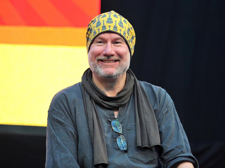 Das Bild zeigt den Comiczeichner Ingo Römling, vom Kopf bis zum Unterbauch. Er lächelt, trägt einen schwarzen Schal und eine gelbe Mütze. 