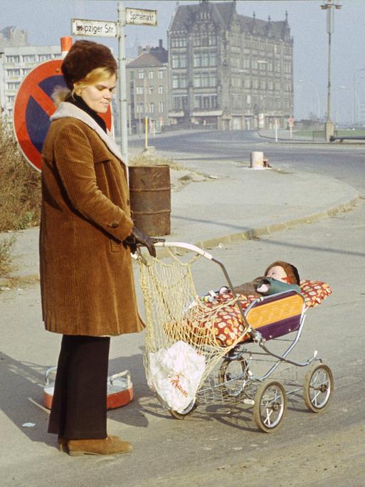 Eine Frau mit Kinderwagen, in dem ein Baby schläft, steht mit nachdenklichem Blick an einer Straßenecke im Berlin der Siebzigerjahre. Die Spuren des Kriegs sind noch im Stadtbild zu erkennen.