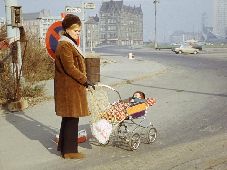 Eine Frau mit Kinderwagen, in dem ein Baby schläft, steht mit nachdenklichem Blick an einer Straßenecke im Berlin der Siebzigerjahre. Die Spuren des Kriegs sind noch im Stadtbild zu erkennen.