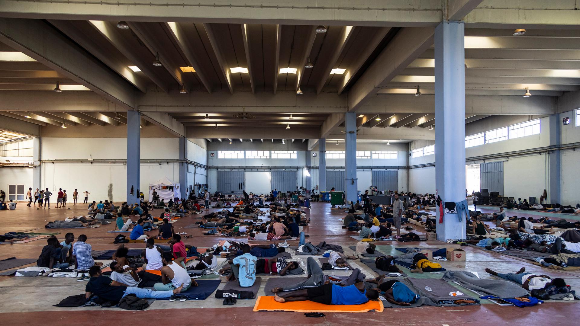 In einer großen Halle lagern Menschen auf Decken und Matten auf dem Boden und sitzen in Gruppen zusammen.