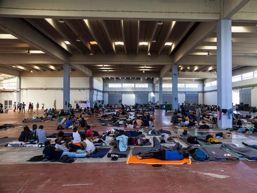 In einer großen Halle lagern Menschen auf Decken und Matten auf dem Boden und sitzen in Gruppen zusammen. In einer großen Halle lagern Menschen auf Decken und Matten auf dem Boden und sitzen in Gruppen zusammen.