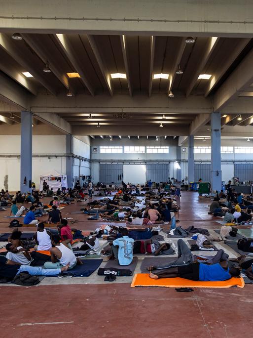 In einer großen Halle lagern Menschen auf Decken und Matten auf dem Boden und sitzen in Gruppen zusammen. In einer großen Halle lagern Menschen auf Decken und Matten auf dem Boden und sitzen in Gruppen zusammen.