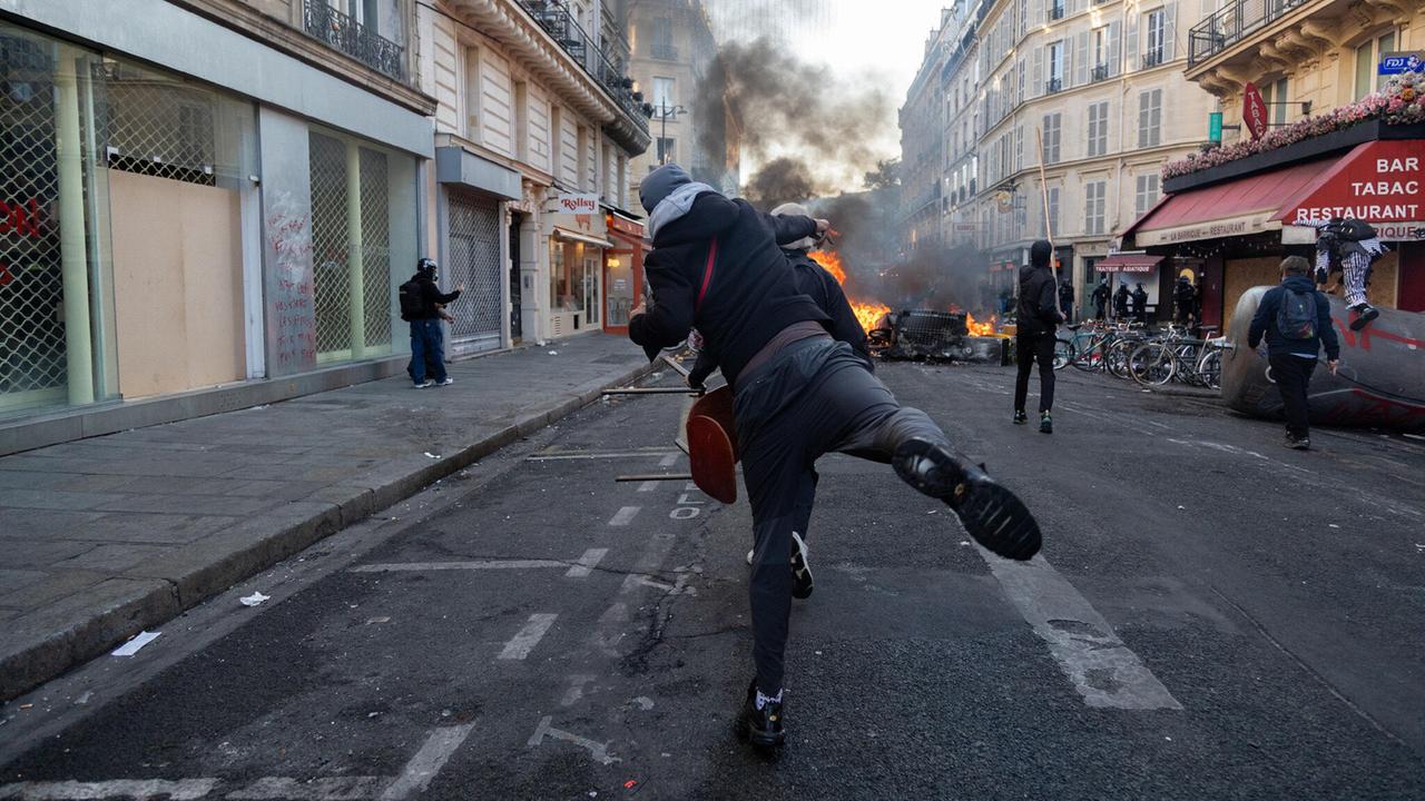 Ein gewalttätiger Demonstrant wirft eine Flasche in Richtung einiger Polizisten hinter einer brennenden Barrikade auf einer Straße in Paris. Ein gewalttätiger Demonstrant wirft eine Flasche in Richtung einiger Polizisten hinter einer brennenden Barrikade auf einer Straße in Paris.