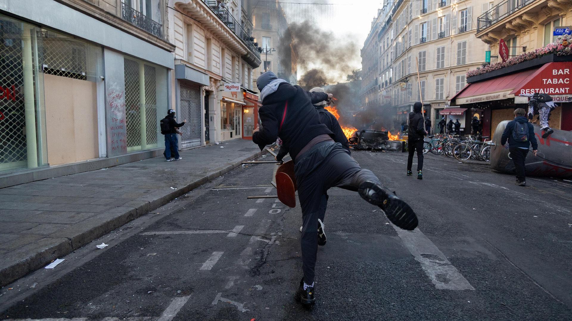 Ein gewalttätiger Demonstrant wirft eine Flasche in Richtung einiger Polizisten hinter einer brennenden Barrikade auf einer Straße in Paris. Ein gewalttätiger Demonstrant wirft eine Flasche in Richtung einiger Polizisten hinter einer brennenden Barrikade auf einer Straße in Paris.