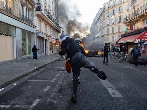 Ein gewalttätiger Demonstrant wirft eine Flasche in Richtung einiger Polizisten hinter einer brennenden Barrikade auf einer Straße in Paris. Ein gewalttätiger Demonstrant wirft eine Flasche in Richtung einiger Polizisten hinter einer brennenden Barrikade auf einer Straße in Paris.
