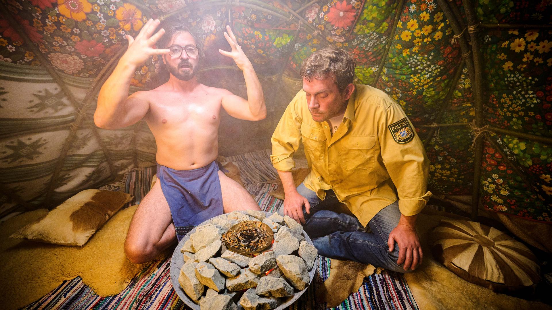 Zwei Schauspieler sitzen in einer Szene des Films "Rehragout-Rendezvous" in einer Art Zelt um ein Feuer. Zwei Schauspieler sitzen in einer Szene des Films "Rehragout-Rendezvous" in einer Art Zelt um ein Feuer.