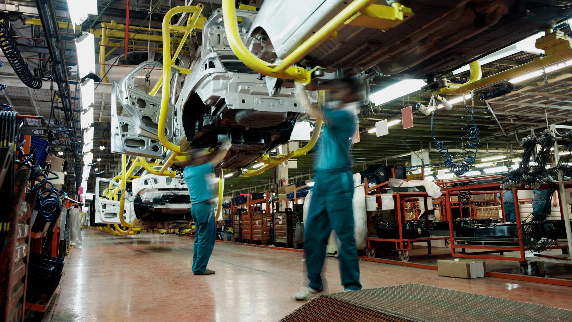 Zwei Arbeiter in grünen Hemden und blauen Hosen arbeiten unter einem Auto in einer Automobilfabrik. Im Hintergrund sind Montagebänder, teilweise fertige Autos, Werkzeuge und Elektrogeräte zu sehen.