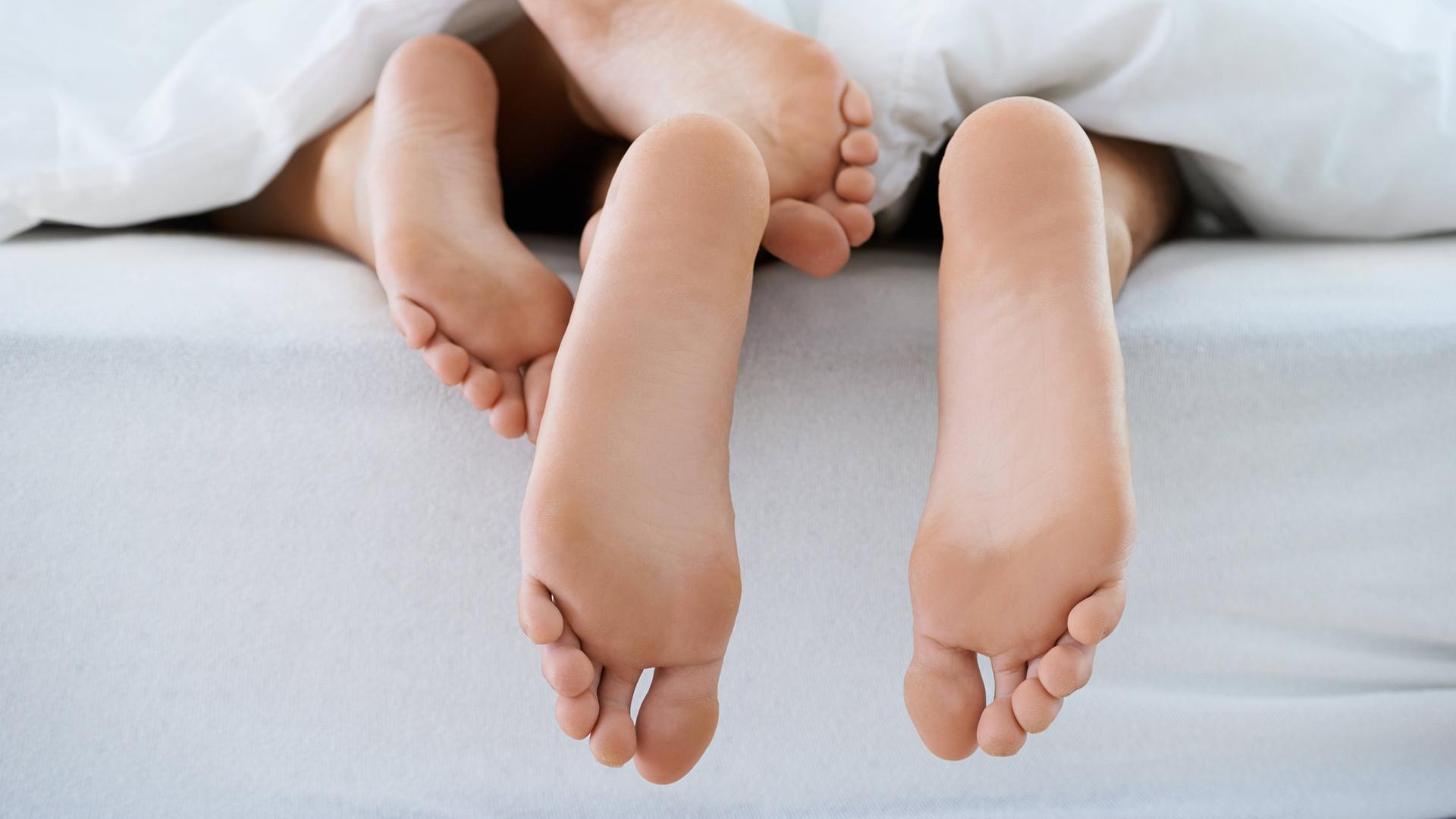 Unter einer Bettdecke schauen zwei Paar Füße hervor. Unter einer Bettdecke schauen zwei Paar Füße hervor.