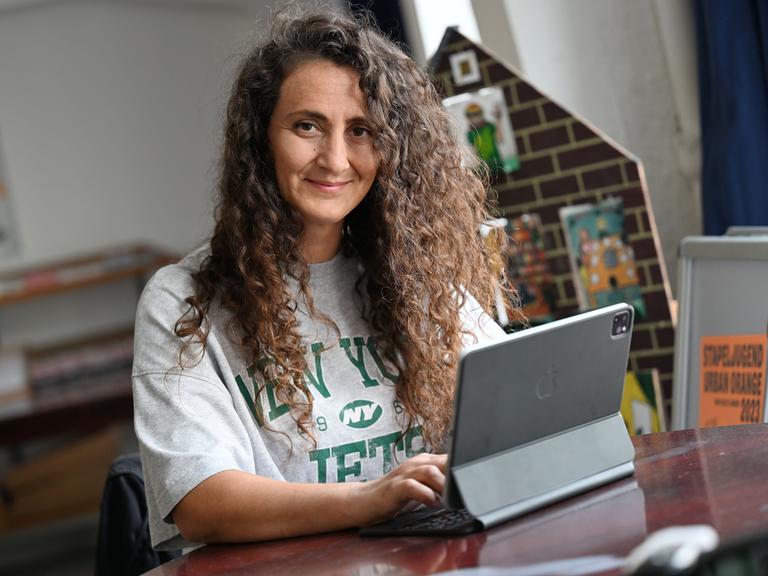 Lale Öztürk, eine Frau mit langen, lockigen Haaren, arbeitet an einem iPad. Sie koordiniert die Sprachförderung der Stadt Duisburg, macht Comedy und liest Menschen ehrenamtlich vor.
