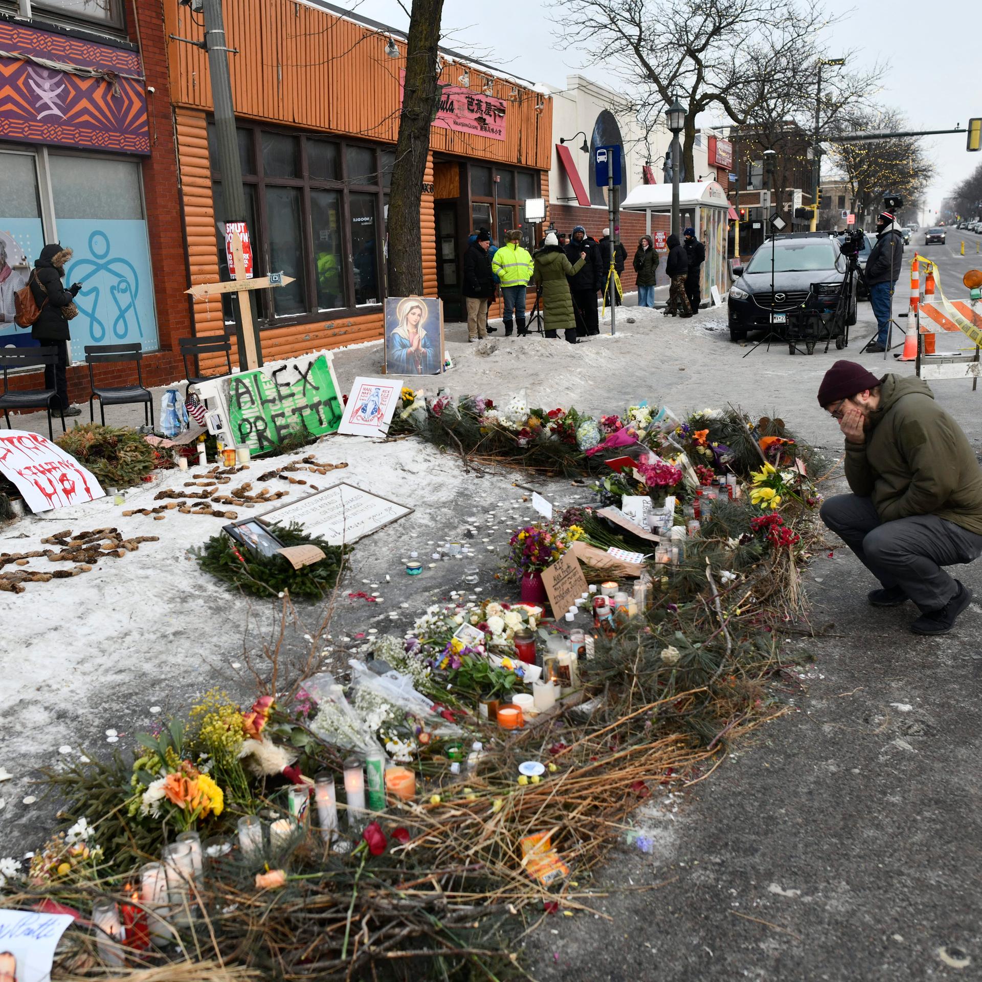 25.01.2026 - Auf dem Boden vor einer Häuserzeile sind Blumen und Inschriften niedergelegt, ein Mann kniet davor, er hält eine Hand vors Gesicht.