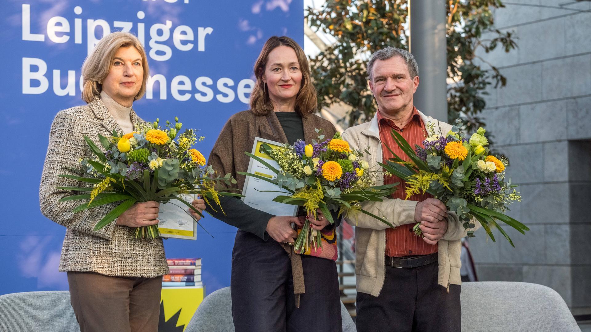 Die drei Buchpreisträger der Leipziger Buchmesse 2026 stehen mit Urkunden und Blumensträußen auf einer Bühne. Die drei Buchpreisträger der Leipziger Buchmesse 2026 stehen mit Urkunden und Blumensträußen auf einer Bühne.