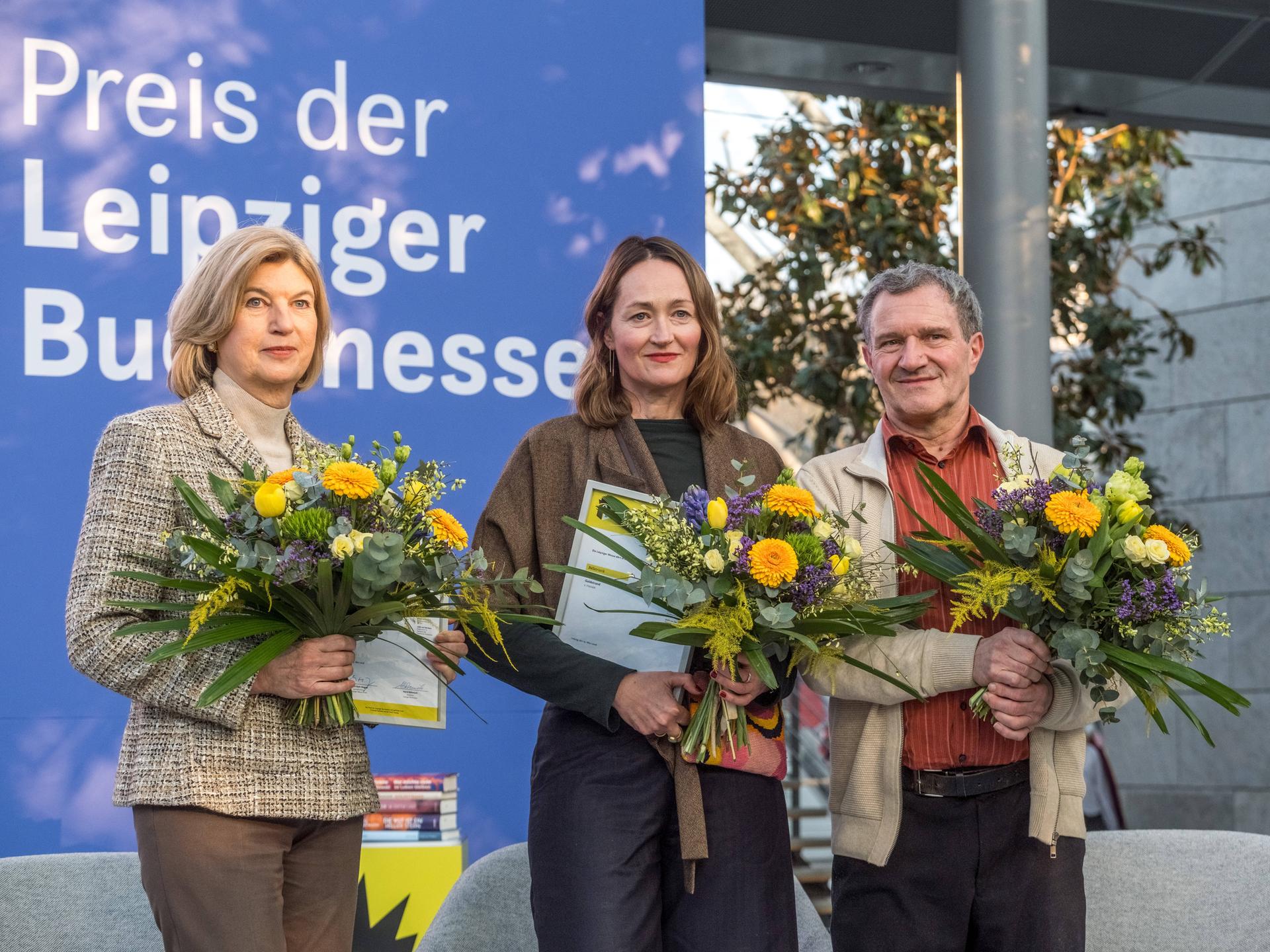 Die drei Buchpreisträger der Leipziger Buchmesse 2026 stehen mit Urkunden und Blumensträußen auf einer Bühne.