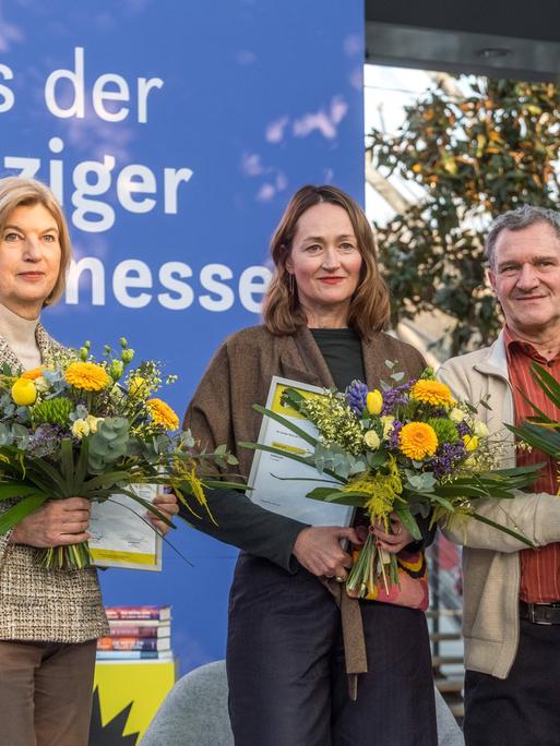Die drei Buchpreisträger der Leipziger Buchmesse 2026 stehen mit Urkunden und Blumensträußen auf einer Bühne. Die drei Buchpreisträger der Leipziger Buchmesse 2026 stehen mit Urkunden und Blumensträußen auf einer Bühne.