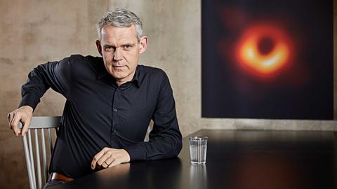 Der Astrophysiker Heino Falcke sitzt auf einem Stuhl, er trägt ein dunkles Hamd und hat graumelierte Haare. Rechts sieht man auf einem Bildschirm einen Feuerball im schwarzen Weltall.