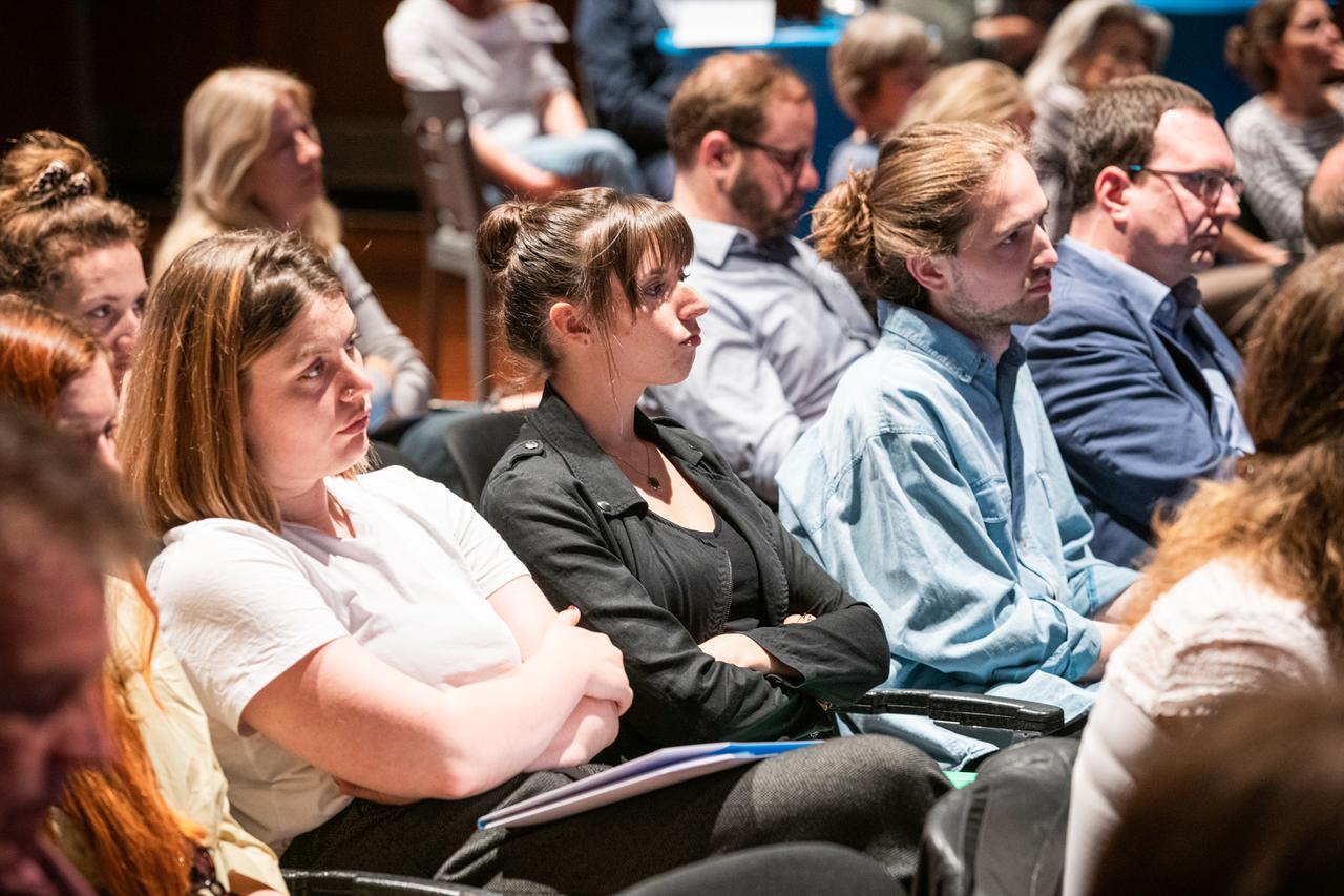 Menschen sitzen in einem Saal und hören einer Diskussion auf der Bühne zu.