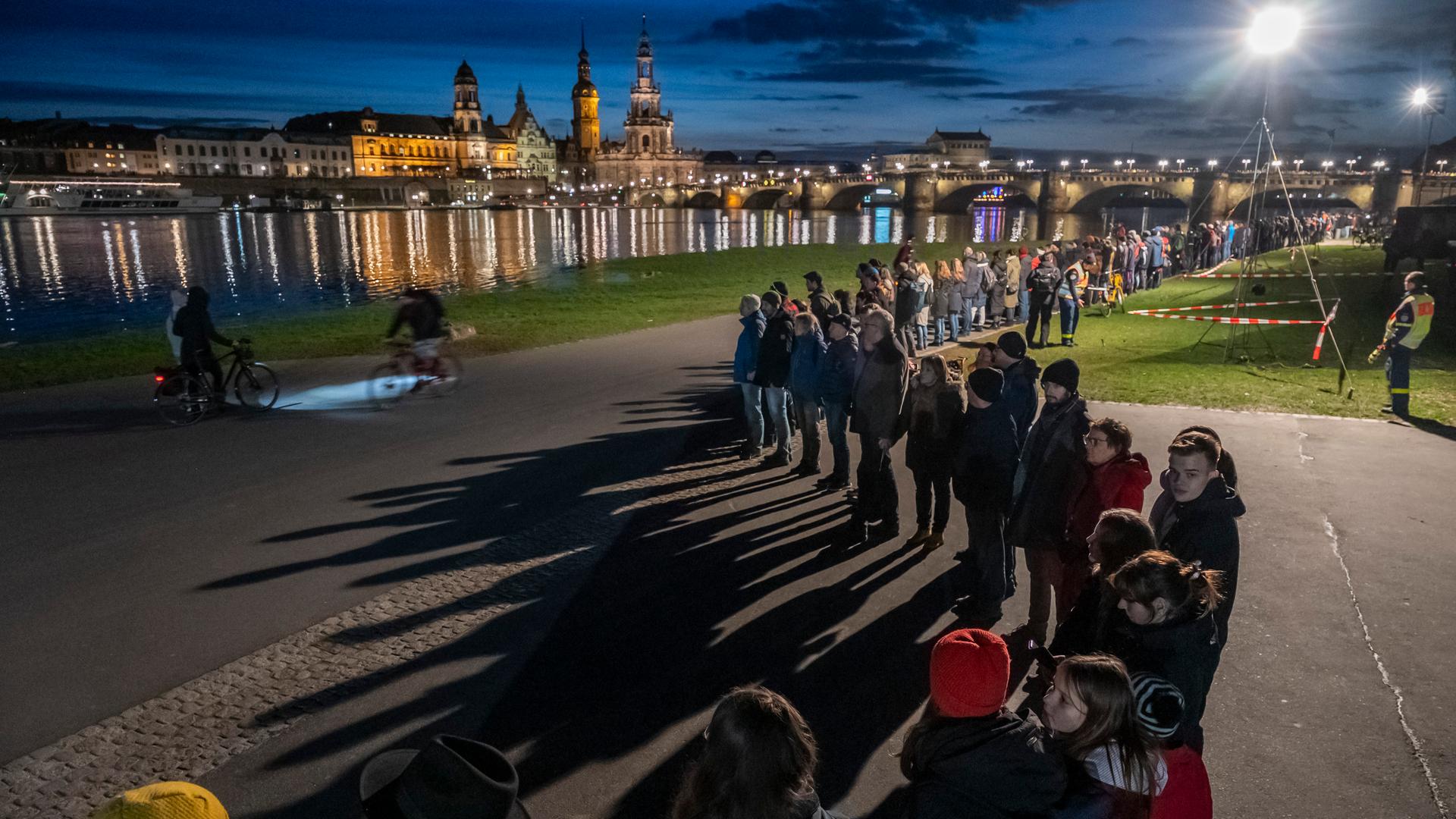 Mehrere tausend Menschen schließen sich am Ufer der Elbe zu einer Menschenkette zusammen. Es ist dunkel. Auf dem Wasser spiegelt sich die Dresdner Altstadt auf der anderen Flusseite. 