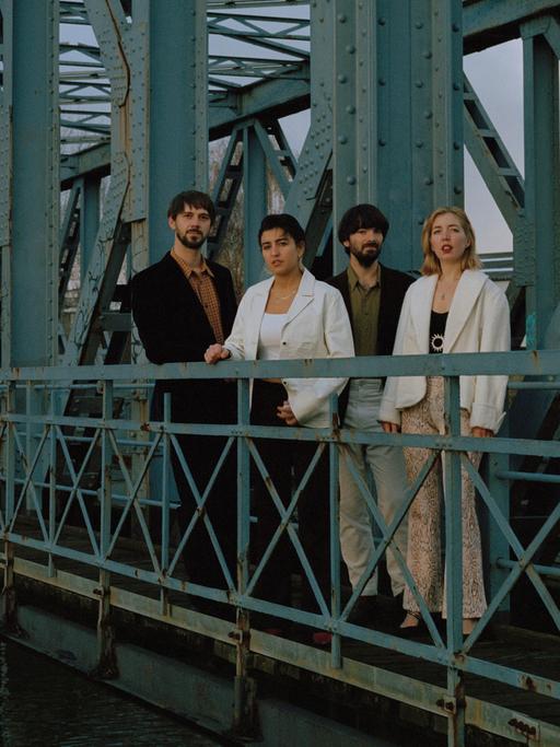 Vier Personen stehen auf einer alten, rostigen Brücke und schauen über die Brüstung in die Kamera.