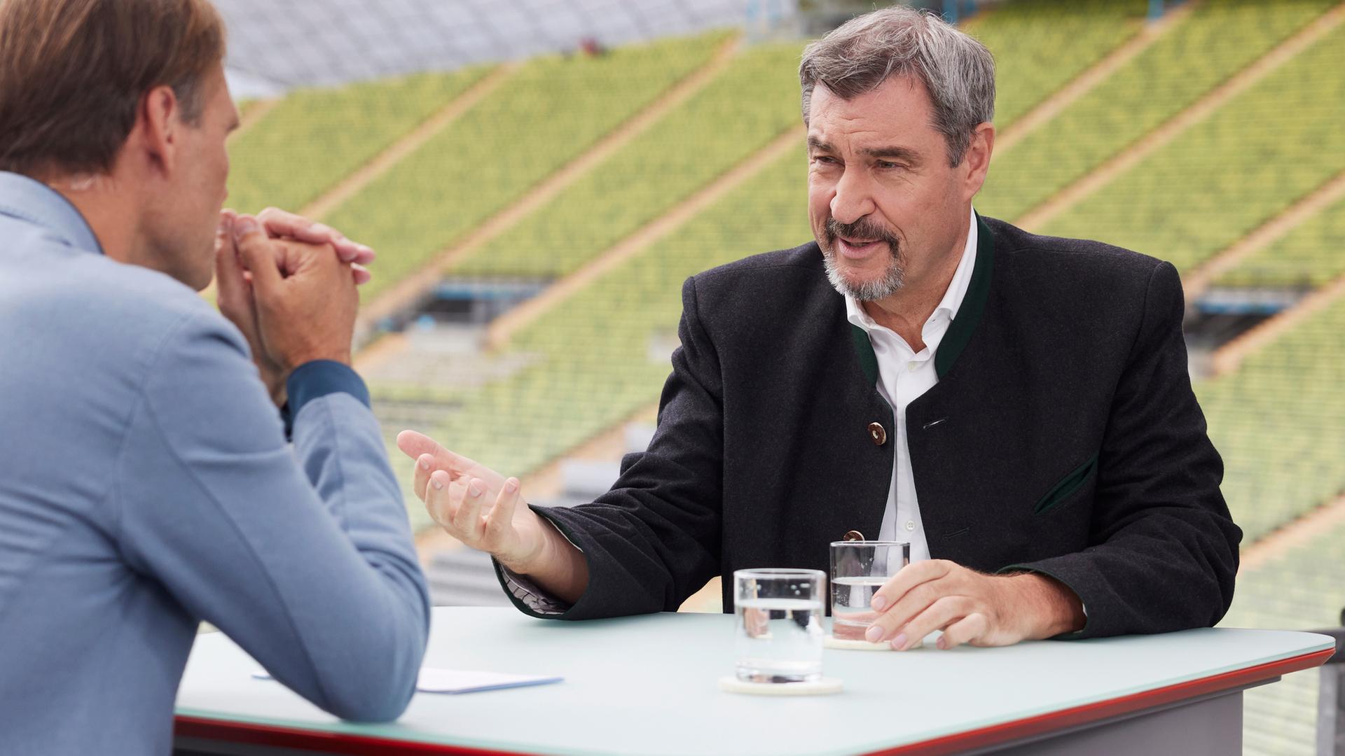 Söder sitzt an einem Tisch mit dem Moderator Wulf Schmiese und sagt etwas. Dahinter sieht man einen Weinberg. 