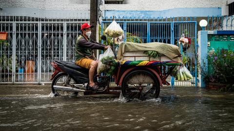 Ein Verkäufer fährt sein mit frischem Gemüse beladenes Motorrad durch eine geflutete Straße.