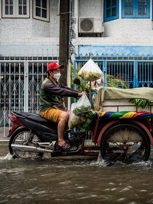 Ein Verkäufer fährt sein mit frischem Gemüse beladenes Motorrad durch eine geflutete Straße.