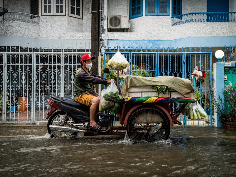 Ein Verkäufer fährt sein mit frischem Gemüse beladenes Motorrad durch eine geflutete Straße.