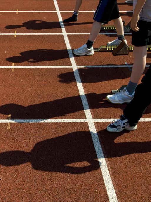 Das Bild zeigt die Beine und Schatten von Kindern, die an der Startlinie einer roten Tartanbahn auf den Beginn eines Rennens warten.