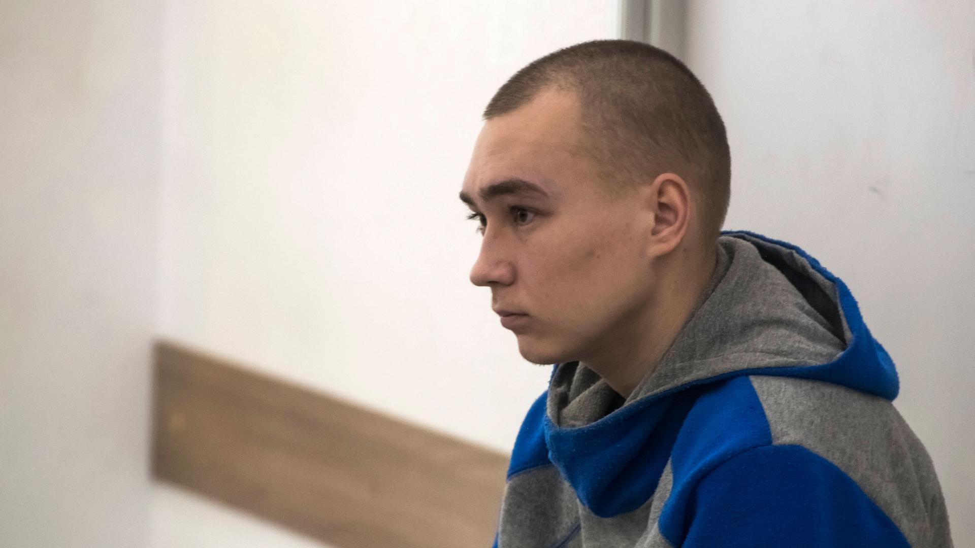 Zu sehen ist  Vadim Schischimarin: Ein junger Mann mit kurzen dunklen Haaren und einer Kapuzenjacke sitzt auf einer Bank in einem kleinen Raum