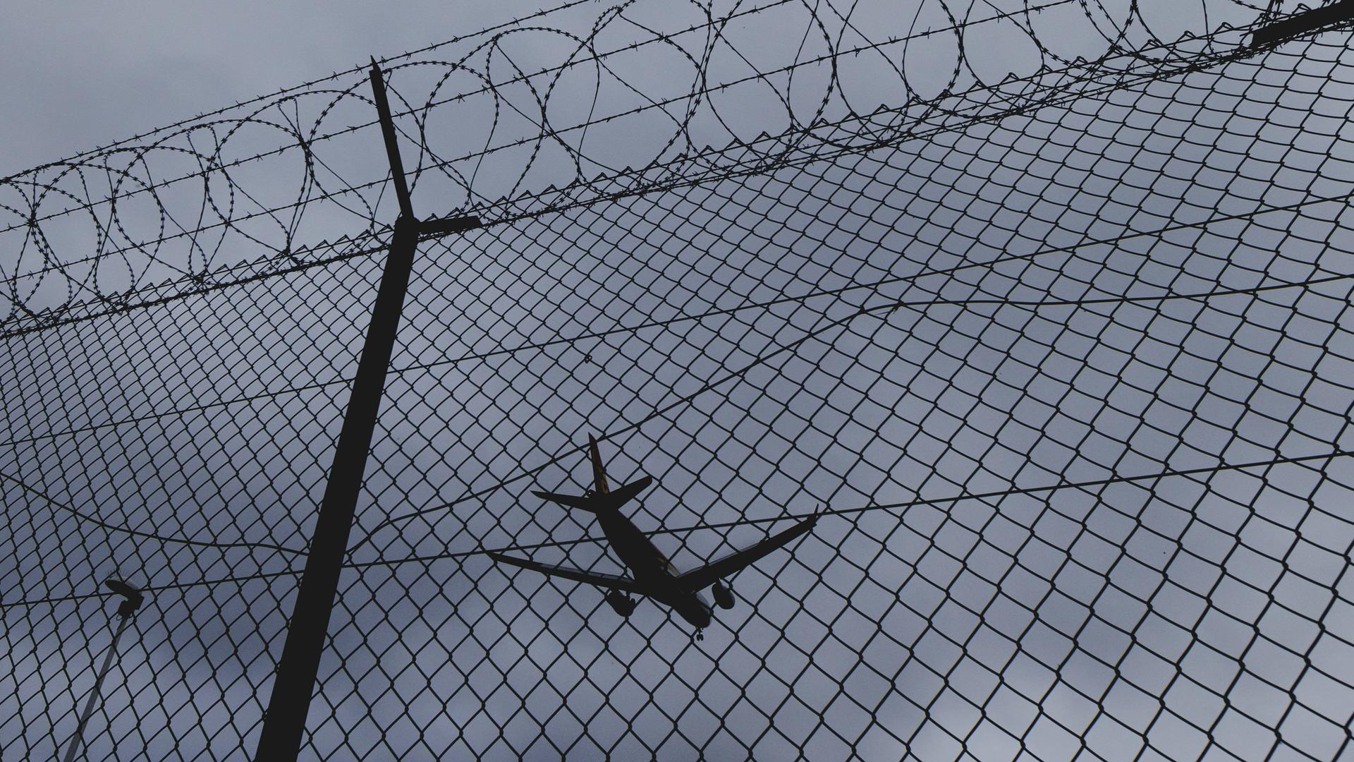 Hinter einem Zaun mit Stacheldraht sieht man ein Flugzeug in der Luft