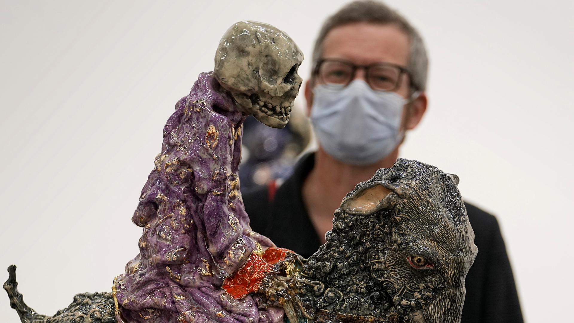 Ein Besucher mit Maske betrachtet auf der Art Cologne eine Arbeit von Carolein Smit aus den Niederlanden. Es ist ein auf einer Ziege reitendes Skelett.