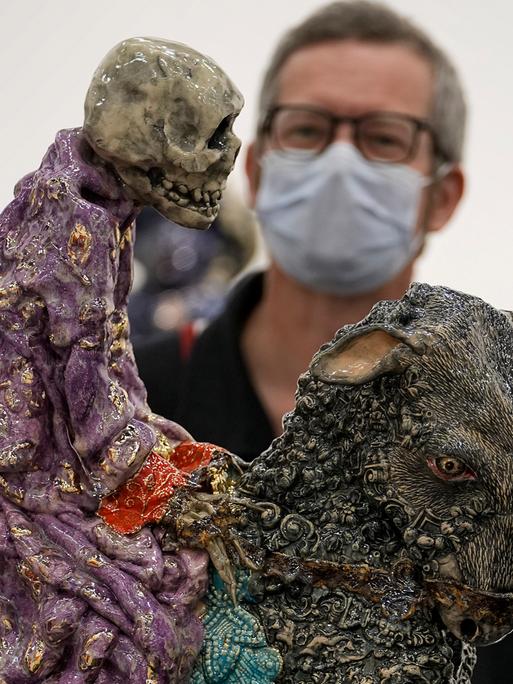 Ein Besucher mit Maske betrachtet auf der Art Cologne eine Arbeit von Carolein Smit aus den Niederlanden. Es ist ein auf einer Ziege reitendes Skelett.