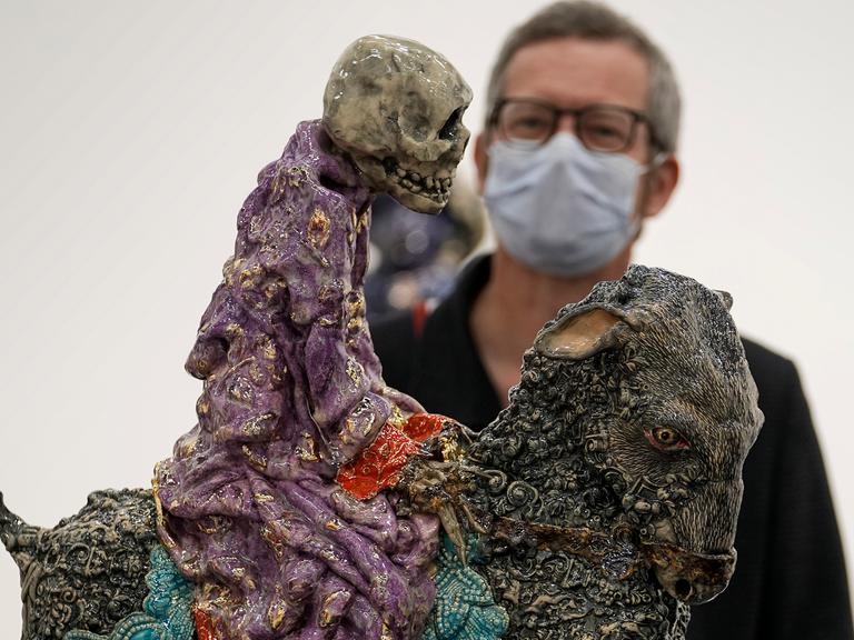 Ein Besucher mit Maske betrachtet auf der Art Cologne eine Arbeit von Carolein Smit aus den Niederlanden. Es ist ein auf einer Ziege reitendes Skelett.