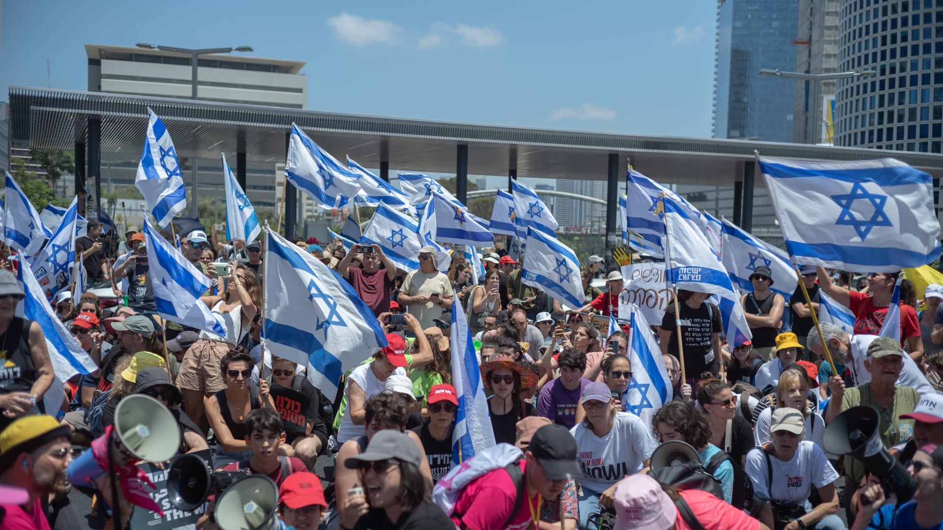 Das Bild zeigt eine große Menschenmenge, die israelische Fahnen schwenkt und eine Straßenkreuzung in Tel Aviv besetzt. Das Bild zeigt eine große Menschenmenge, die israelische Fahnen schwenkt und eine Straßenkreuzung in Tel Aviv besetzt.