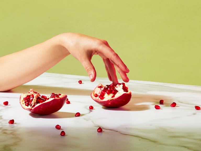 Ein geöffneter Granatapfel liegt auf einem Tisch. Einzelne Kerne sind verstreut. Eine Hand berührt sanft einen Teil des Granatapfels. Das Bild mutet sinnlich an.