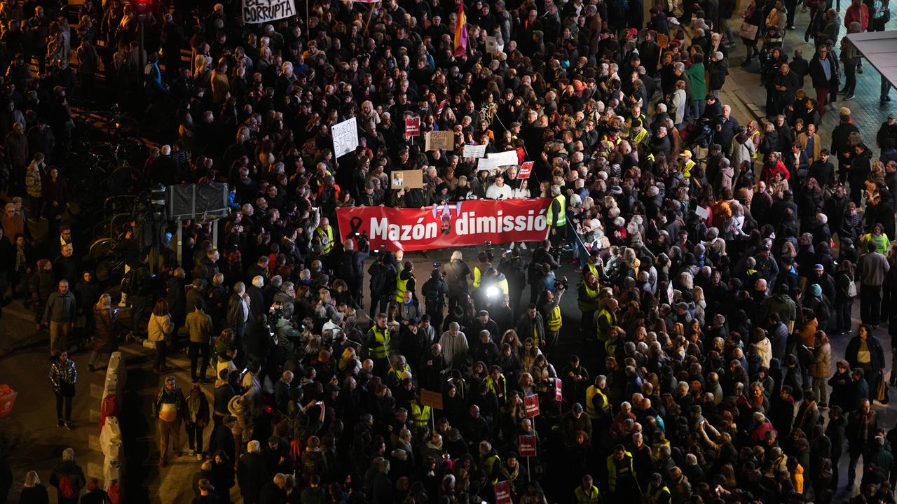 Viele Menschen protestieren im Dunkeln auf einer weitläufigen Straße. Manche von ihnen tragen Transparente. Medienvertreterinnen und Medienvertreter filmen und machen Fotos. Ihre Blitzlichter und Beleuchtungsgeräte erhellen die Szenerie.