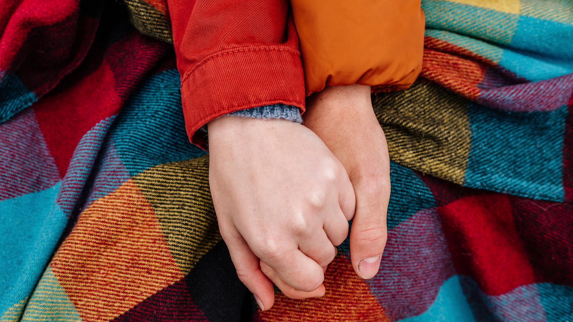 Ein Junge und ein Mädchen halten Händchen auf einer karierten Decke. In der Großaufnahme sieht man nur die Hände der beiden Teenager. 